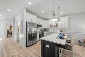 A wide view of an open-concept living area shows a kitchen with a dark island and white cabinets. The space has light wood-look flooring and opens into a dining and living area, with a bedroom visible down a hallway.