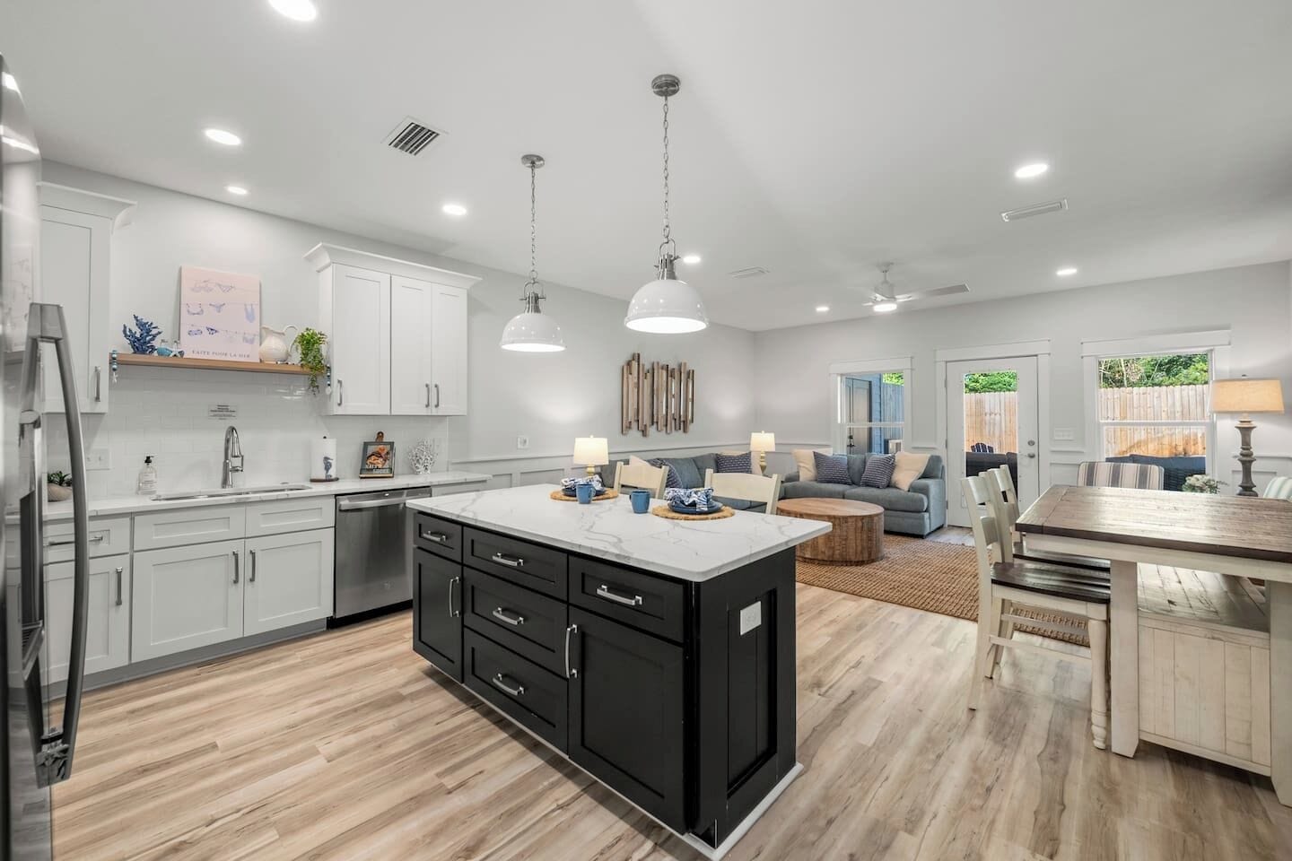 An open-concept living area features a kitchen with a large dark gray island and white cabinets. The space extends into a dining area and living room with a gray sectional sofa, all on light wood-look flooring.