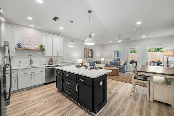 An open-concept living area features a kitchen with a large dark gray island and white cabinets. The space extends into a dining area and living room with a gray sectional sofa, all on light wood-look flooring.