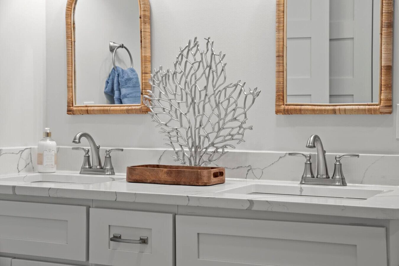 A bathroom double vanity has a white marble countertop with two undermount sinks and brushed nickel faucets. A silver coral sculpture is centered between the sinks, and two mirrors with braided wood frames hang on the wall.