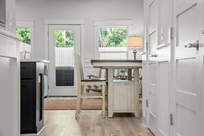 A short hallway with white doors leads into a bright room with a rustic dining table. A glass door and a window at the back of the room look out onto a fenced-in yard. - Towne Treasures B ~ Brand-New Luxe Coastal Retreat with Private Cocktail Pool (Panama City Beach)