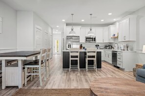 A spacious open-concept kitchen and dining area with light wood-look flooring is shown. The kitchen has a large dark island, white cabinets, and stainless steel appliances. A long dining table with a bench and chairs is visible to the left.v - Towne Treasures B ~ Brand-New Luxe Coastal Retreat with Private Cocktail Pool (Panama City Beach)