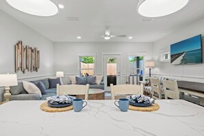 A view from a kitchen island shows an open-concept living area. In the foreground, two place settings with blue dishes sit on the white countertop. Beyond the counter is a dining area and a living room with a large blue sectional sofa and a flatscreen TV