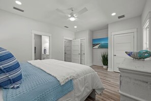 A clean, bright bedroom with light wood-look flooring features a bed with a white comforter and blue blanket. A white dresser with a TV displaying a beach scene is on the right, and a bathroom is visible through an open door to the left. - Towne Treasures B ~ Brand-New Luxe Coastal Retreat with Private Cocktail Pool (Panama City Beach)