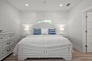 A clean, modern bedroom is shown with a distressed white wooden bed frame and a white paneled accent wall. The bed is covered with a white comforter and blue and white striped pillows. Matching distressed nightstands with lamps flank the bed.