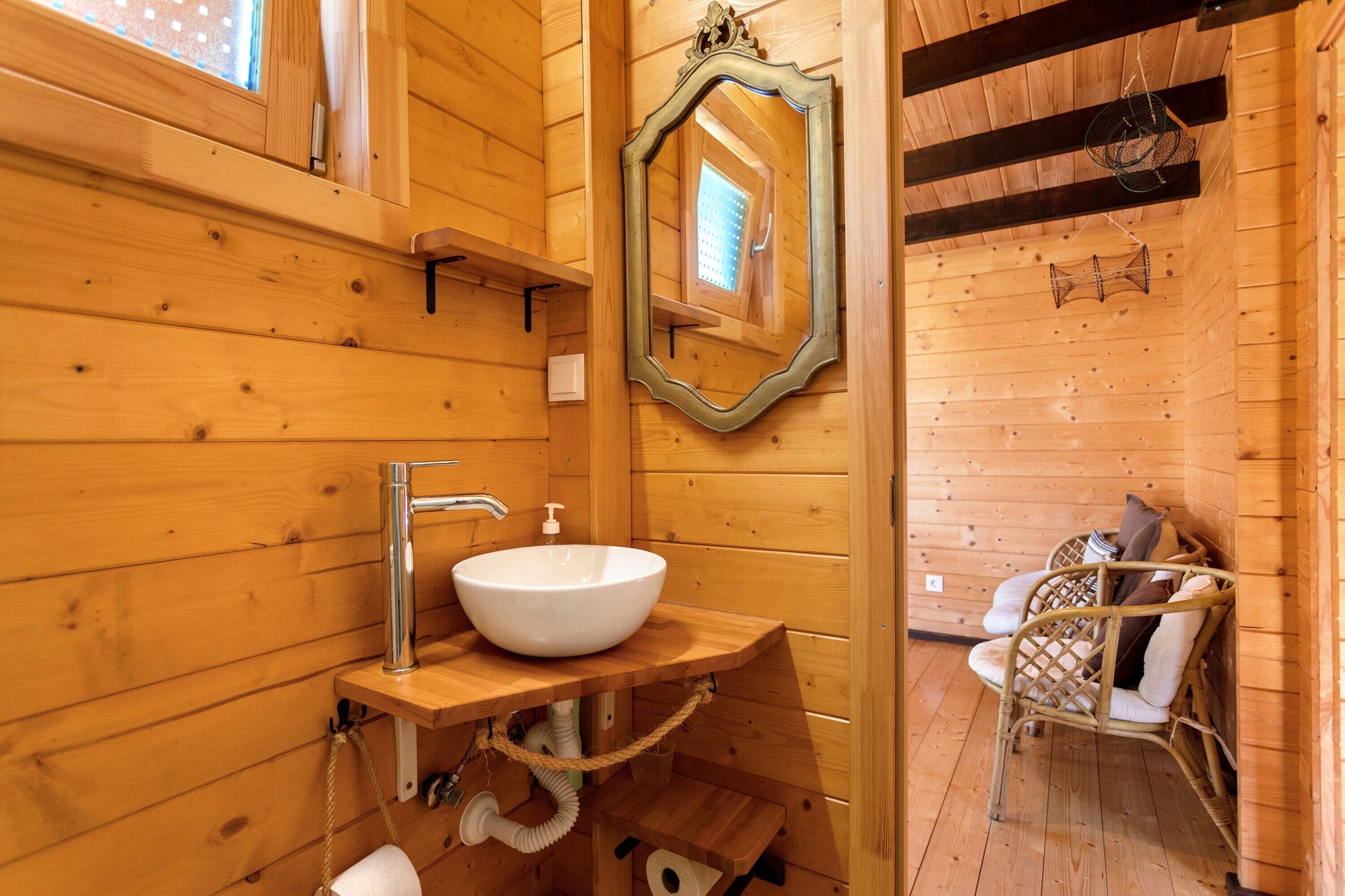A wooden cabin bathroom with a white sink, wooden shelves, a mirror, and a wicker chair.