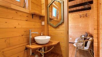 A wooden cabin bathroom with a white sink, wooden shelves, a mirror, and a wicker chair.