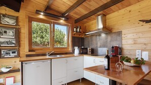 A modern kitchen with wooden cabinetry, a built-in dishwasher, a microwave, and a large window with a view of greenery. - Cabin 'Pátio do Pescador' with Private Terrace, Private Garden, and Wi-Fi (Peniche)