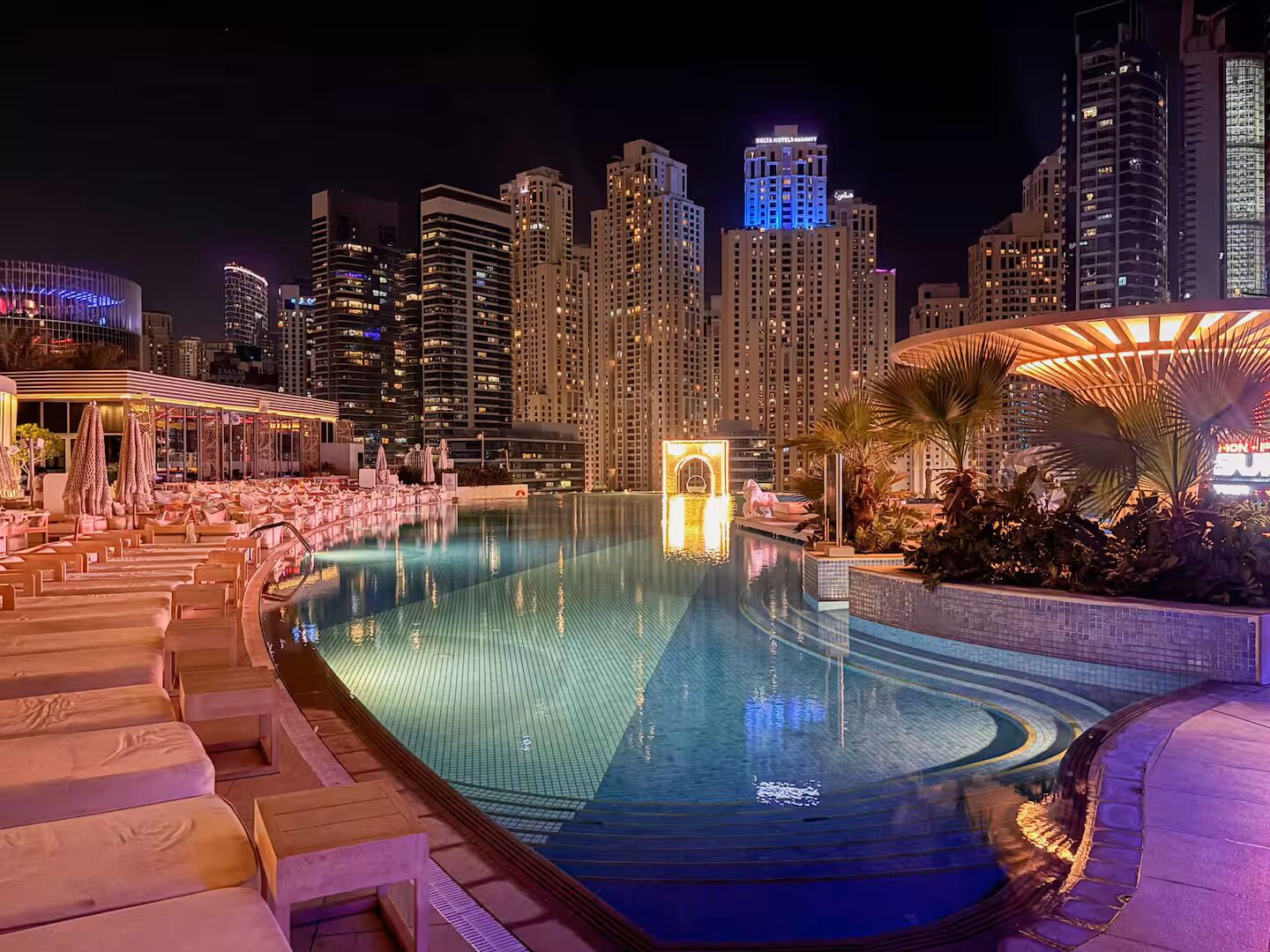 Vue nocturne d’une piscine d’hôtel, avec des chaises longues et, en arrière-plan, le profil de la ville illuminée.