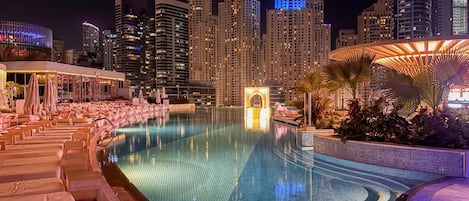 A nighttime view of a hotel pool with lounge chairs and a city skyline in the background.