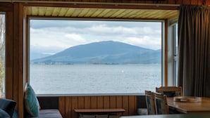 Une cabane dotée d’une grande fenêtre donnant sur un lac et des montagnes. À l’intérieur, on trouve une table en bois et un banc rembourré.