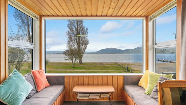 Un chalet confortable doté d’une grande fenêtre donnant sur un lac et des montagnes. À l’intérieur, on trouve un banc en bois et un banc rembourré agrémenté de coussins colorés.
