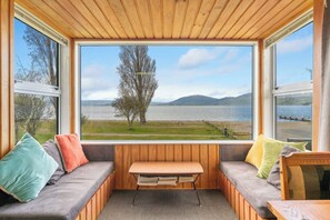 Een knus huisje met een groot raam dat uitzicht geeft op een meer en bergen. Binnenin staat een houten bank en een comfortabele bank met kleurrijke kussens.
