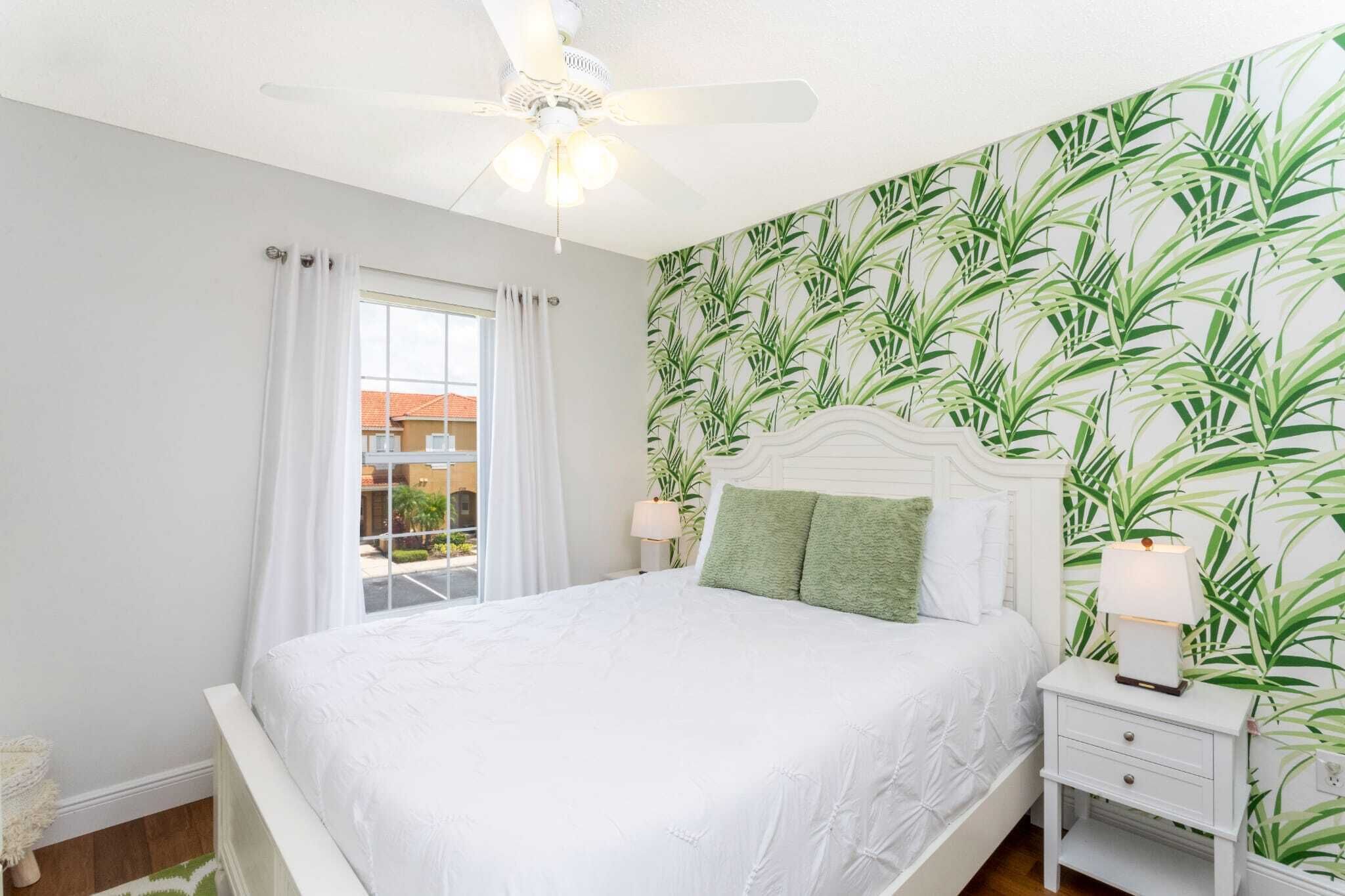 Une chambre à coucher avec un lit, des rideaux blancs, un ventilateur de plafond, une table de chevet avec une lampe et un mur orné d’un motif de feuilles vertes.