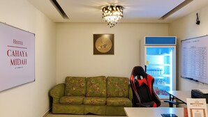 Habitación de hotel con un sofá floral verde, una silla gamer, un escritorio con un teléfono inteligente, un cuadro enmarcado y una pantalla mural que muestra los gastos del hotel.