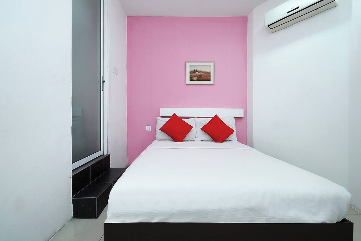 Een compacte, netjes ingerichte slaapkamer met een wit bed, roze wanden en één rode kussen.
