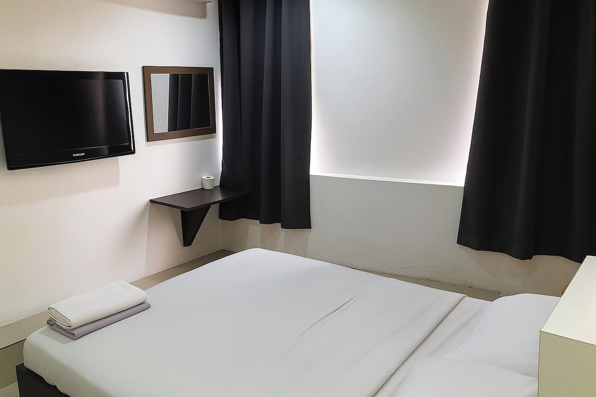 Een hotelkamer met een bed, een flatscreen televisie aan de muur, een klein nachtkastje en een raam met donkere gordijnen.