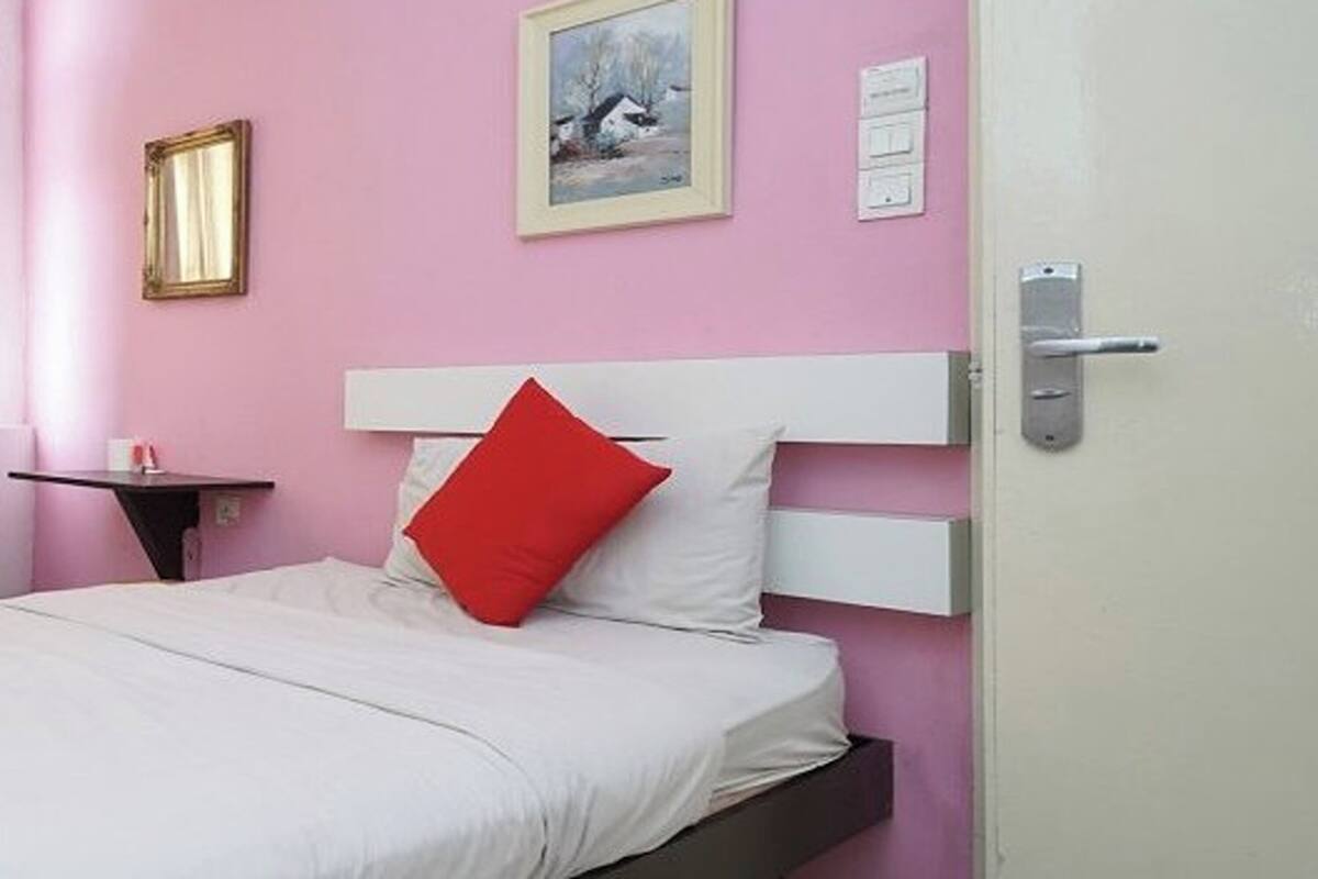 Een slaapkamer met een roze muur, een wit bed, een rood kussen en een lijst met een afbeelding.
