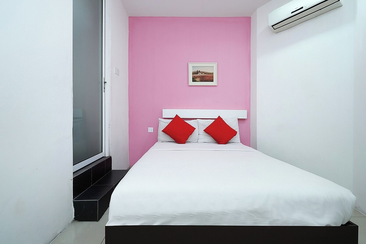Une petite chambre à coucher, bien rangée, avec un lit blanc, des murs roses et un seul coussin rouge.