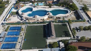 Ett flygbildsfoto av ett semesterområde med flera pooler, en sportplan och en parkeringsplats.