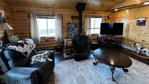 A cozy cabin interior with a wooden ceiling, a fireplace, a television, and a dog resting on a leather couch. - Log Cabin in Whitehall (Whitehall)