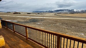 A wooden deck with a metal railing overlooking a rural landscape with mountains and sparse vegetation. - Log Cabin in Whitehall (Whitehall)