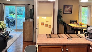 Una cocina con un mueble de madera, una cafetera y una mesa de comedor con sillas. Hay una parrilla fuera de la puerta corrediza de vidrio.