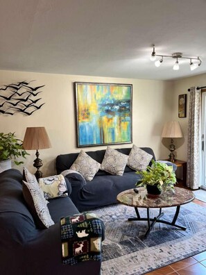 A living room with a dark sofa, patterned cushions, a large abstract painting, a lamp, a coffee table with a plant, and a decorative wall piece.