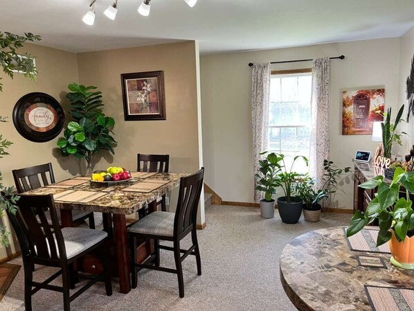 A dining area with a round table, chairs, potted plants, framed artwork, and a window with curtains.