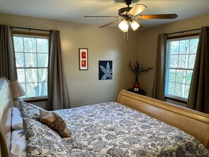 A bedroom with a bed, a ceiling fan, two windows with curtains, and wall art.