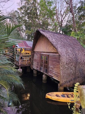 Una casa flotante de madera con techo de paja, sostenida por pilotes en un cuerpo de agua, rodeada de exuberante follaje.