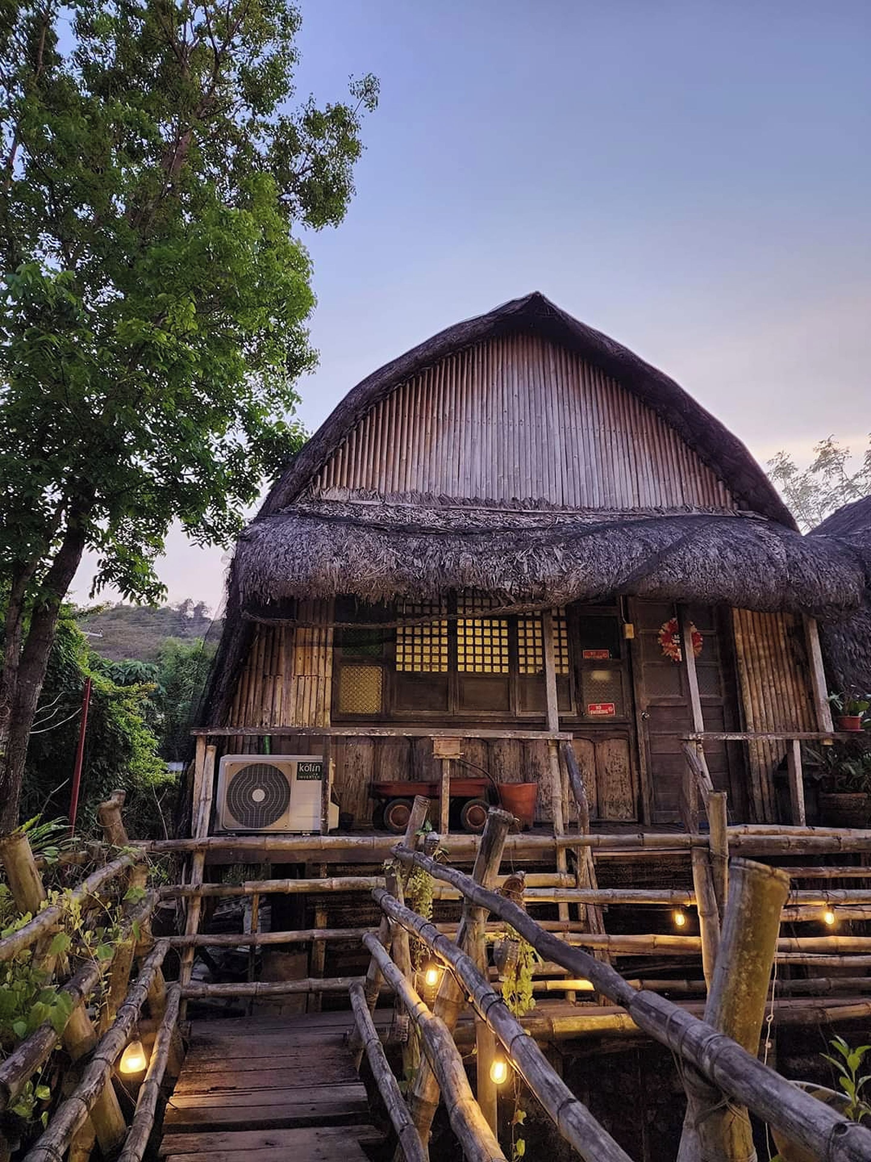 A rustic wooden house with a thatched roof, a wooden porch, and a wooden staircase leading to the entrance.