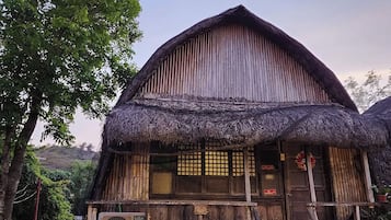 A rustic wooden house with a thatched roof, a wooden porch, and a wooden staircase leading to the entrance.