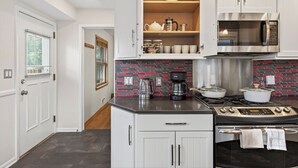 A modern kitchen with white cabinetry, a black countertop, a red and black tiled backsplash, a stainless steel microwave, a stove, and a coffee maker.