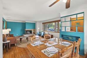 Un coin repas avec une table en bois dressée pour quatre personnes, une cheminée et une vue sur l’extérieur grâce à de grandes fenêtres.