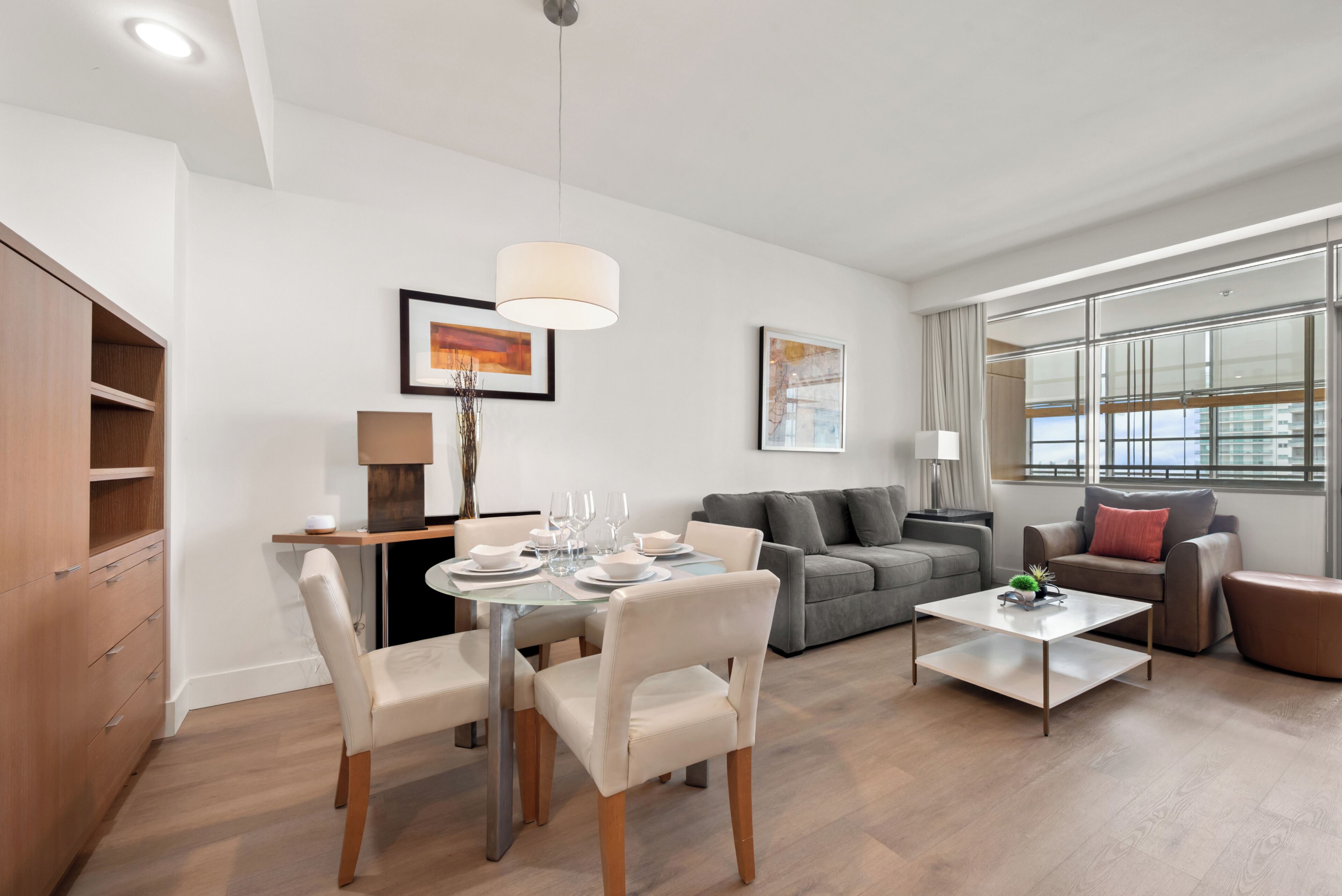 A modern living room with a dining area, a sofa set, and a large window providing natural light.