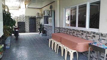 Un espacio exterior cubierto para comer con una larga mesa y varias sillas, una motocicleta estacionada a un lado, y una ventana con persianas.