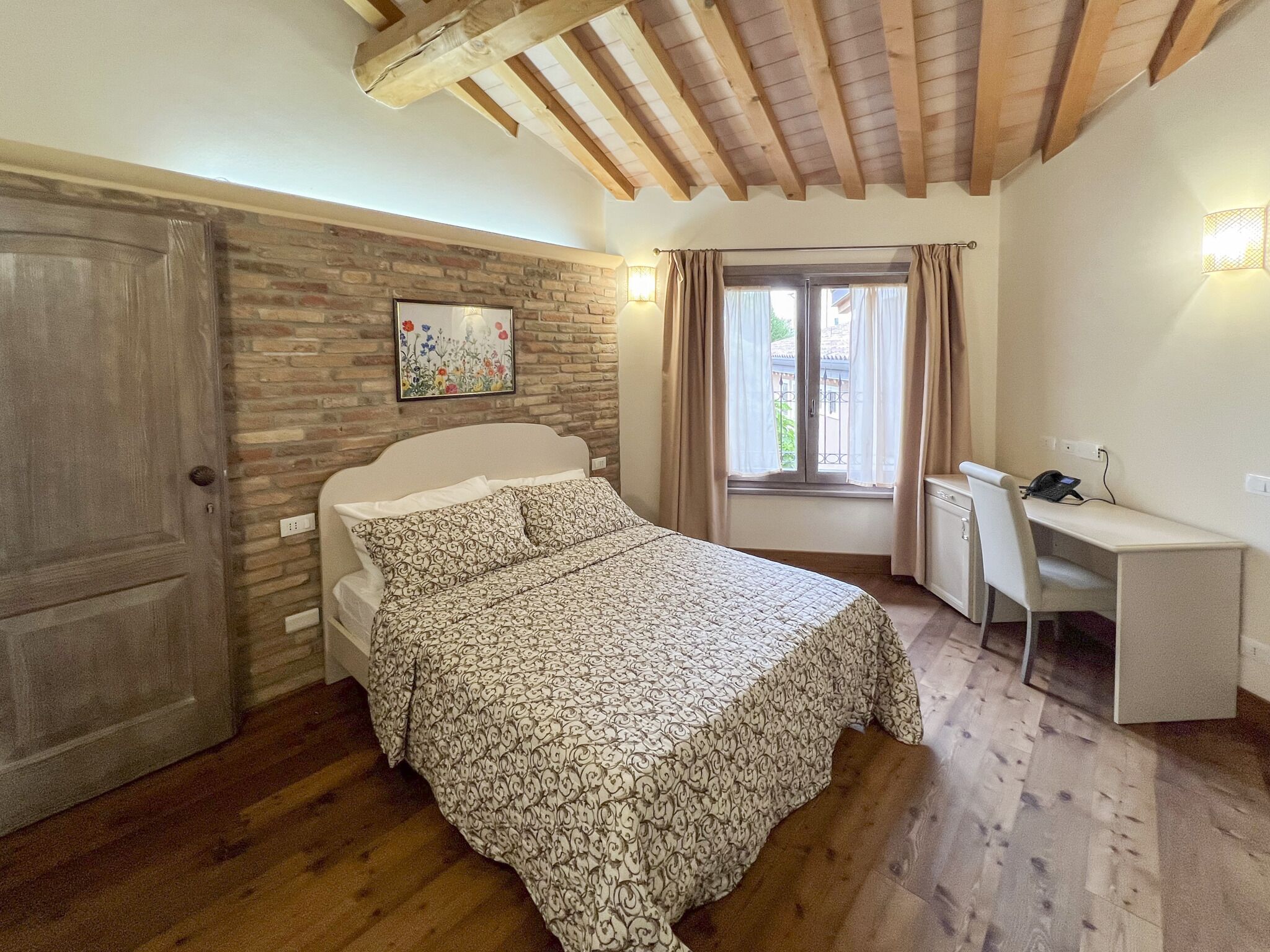 Ein Schlafzimmer mit Bett, Schreibtisch, Computer, einem Fenster mit Vorhängen, einer Backsteinwand und einer Tür.