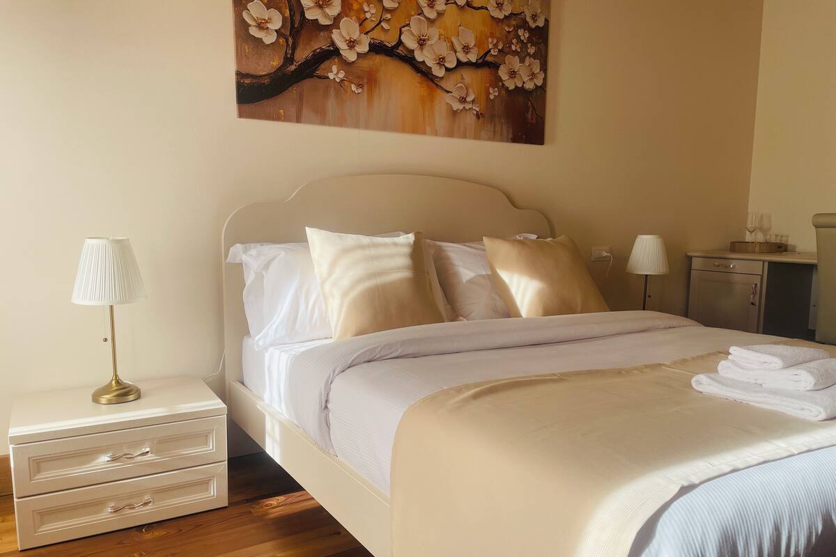 Una cama bien hecha con s ábanas blancas y una toalla doblada al lado. Hay una mesita de noche con una lámpara y un cajón. Un cuadro de flores cuelga en la pared sobre la cama.