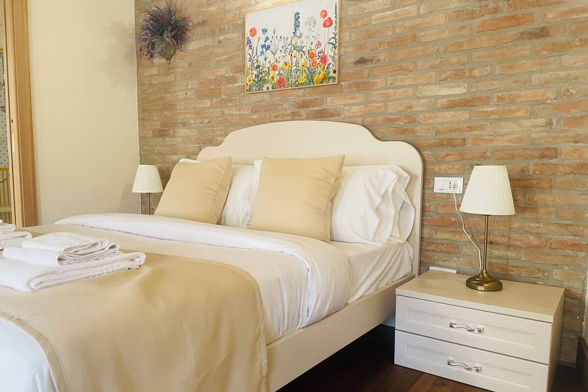 Una cama bien hecha con sábanas blancas y almohadas color beige, una lámpara de noche y una mesita de noche pequeña de color blanco con cajones.