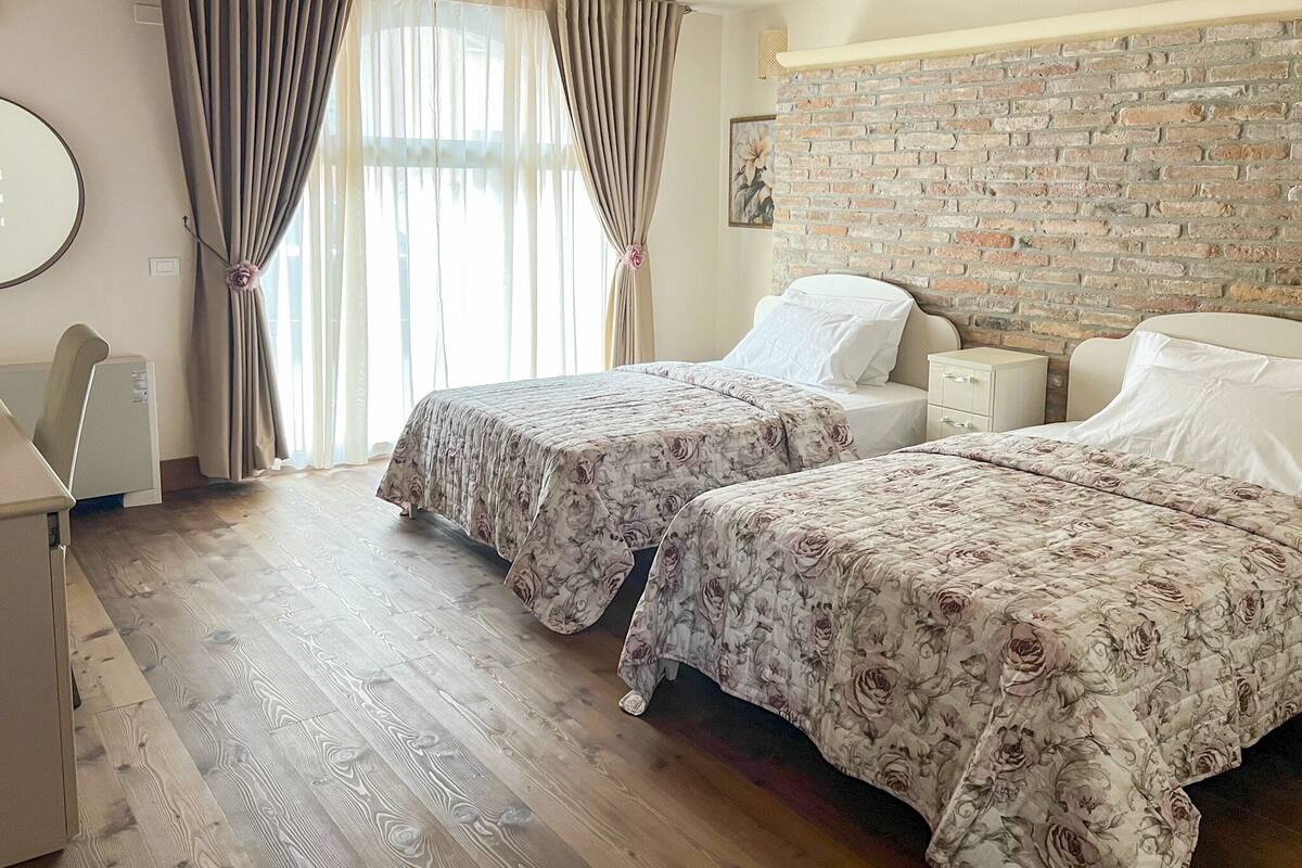 Habitación de hotel con dos camas, pared de ladrillo, suelo de madera y un ventanal con cortinas.