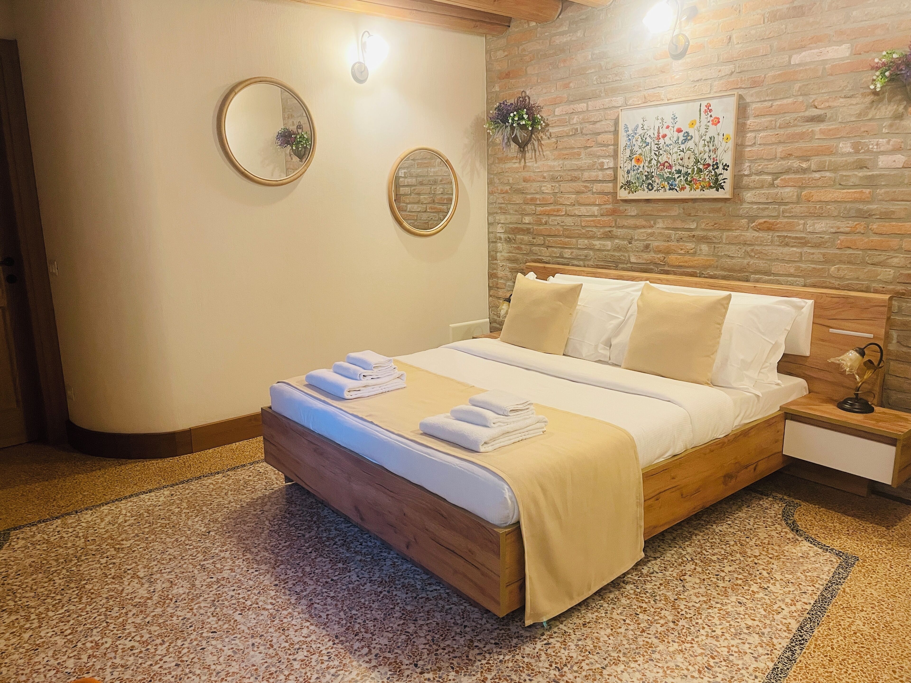 Un letto rifatto con lenzuola bianche e asciugamani piegati, una testiera in legno, un comodino con una lampada e un muro in mattoni con un dipinto floreale.