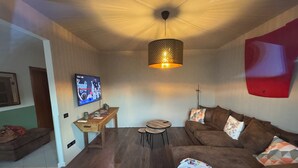 Een gezellige woonkamer met een bruine bank, een salontafel, een aan de muur bevestigde televisie en een hanglamp.