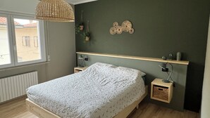 Een slaapkamer met een houten bed, een hanglamp, een wandplank met decoratieve spullen, en een raam met uitzicht op gebouwen.
