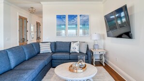 Ein Wohnzimmer mit einem blauen Ecksofa, einem runden Couchtisch mit Dekoration, einem Flachbildfernseher und gerahmten Kunstwerken an der Wand.