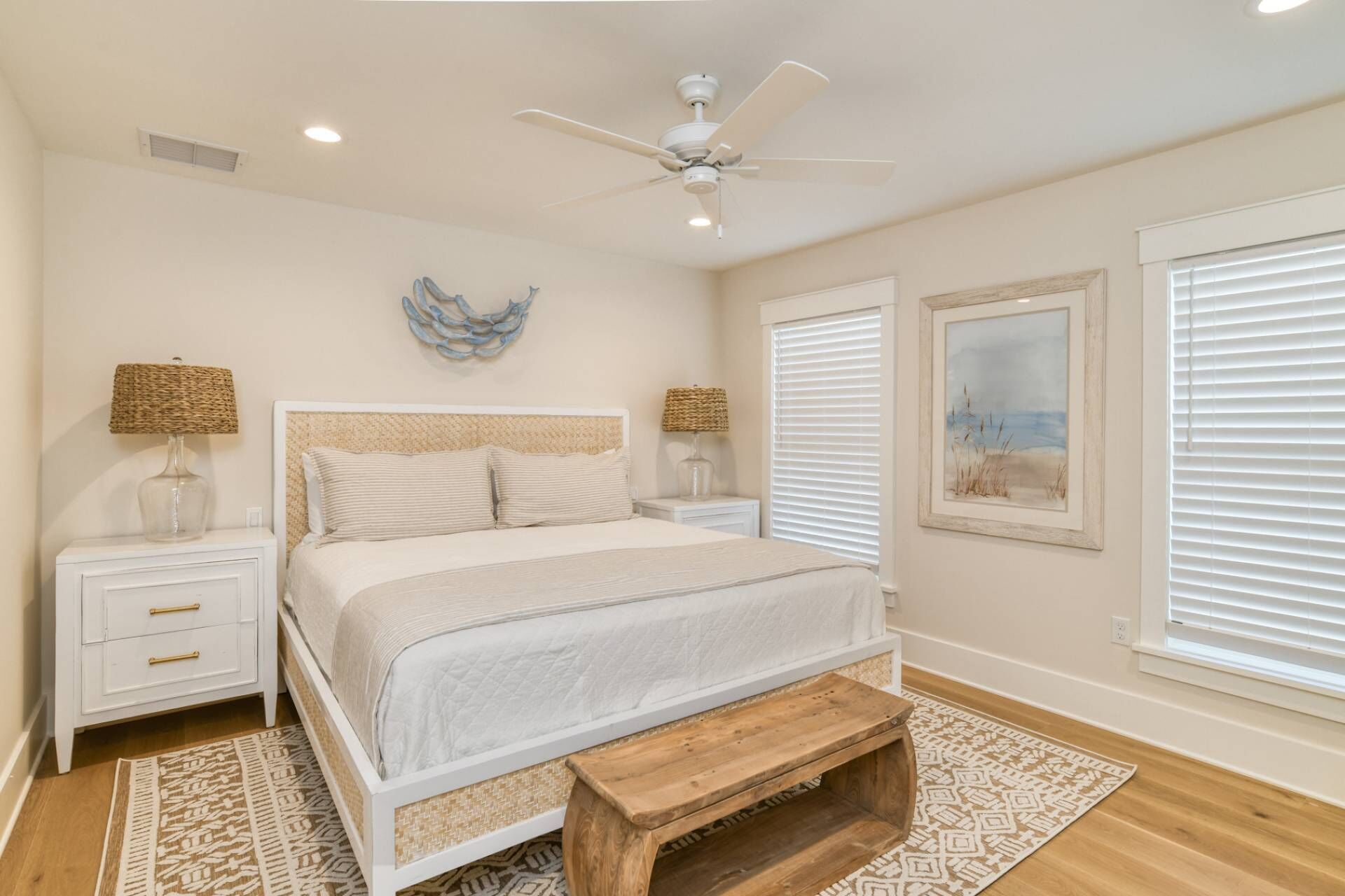 A bedroom with a bed, nightstands, a ceiling fan, a decorative wall piece, a window with blinds, and a framed picture on the wall.