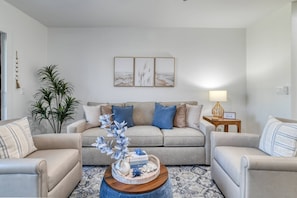 Una sala de estar con un sofá color beige, dos sillones, una mesa de centro con objetos decorativos, una mesita de noche con una lámpara y adornos en la pared.
