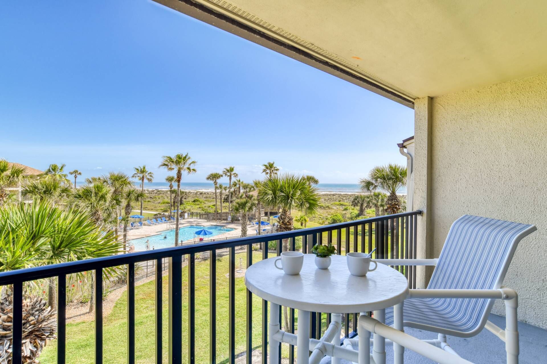 A balcony with a view of a pool and beach, a table with cups and a plant, and a striped chair.