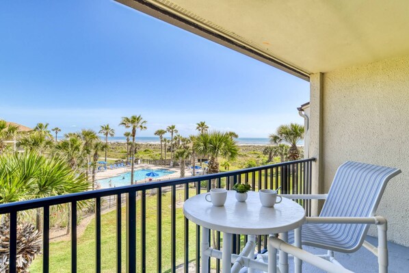 A balcony with a view of a pool and beach, a table with cups and a plant, and a striped chair.