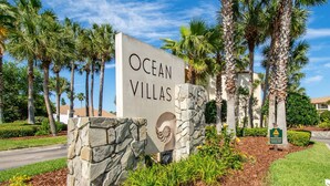 Een bordje voor Ocean Villas, met een stenen voetstuk en een logo, omringd door palmbomen en een aangelegde tuin.